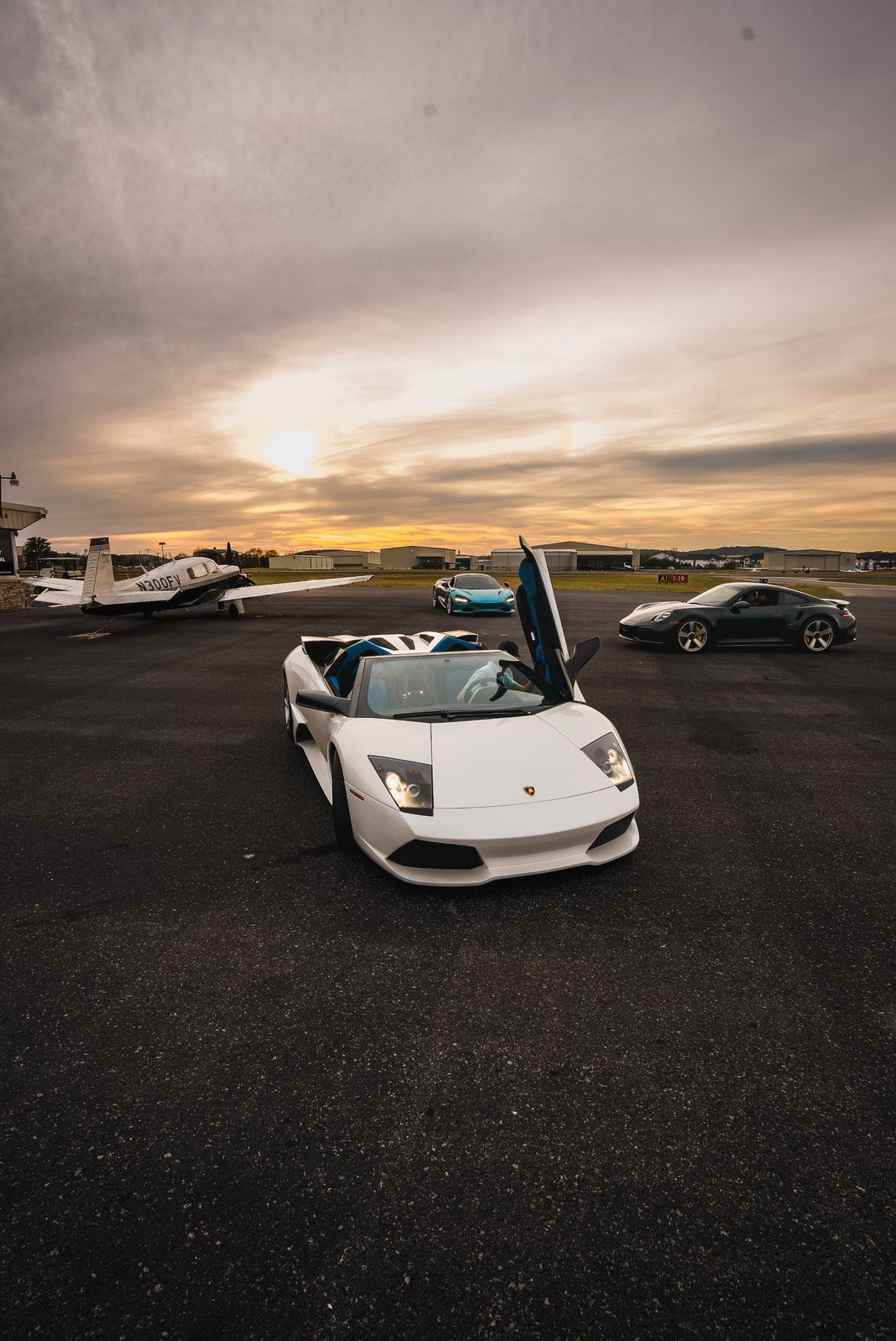 White Lamborghini Murcielago Roadster with a McLaren, Porsche and private plane at an airfield at sunset