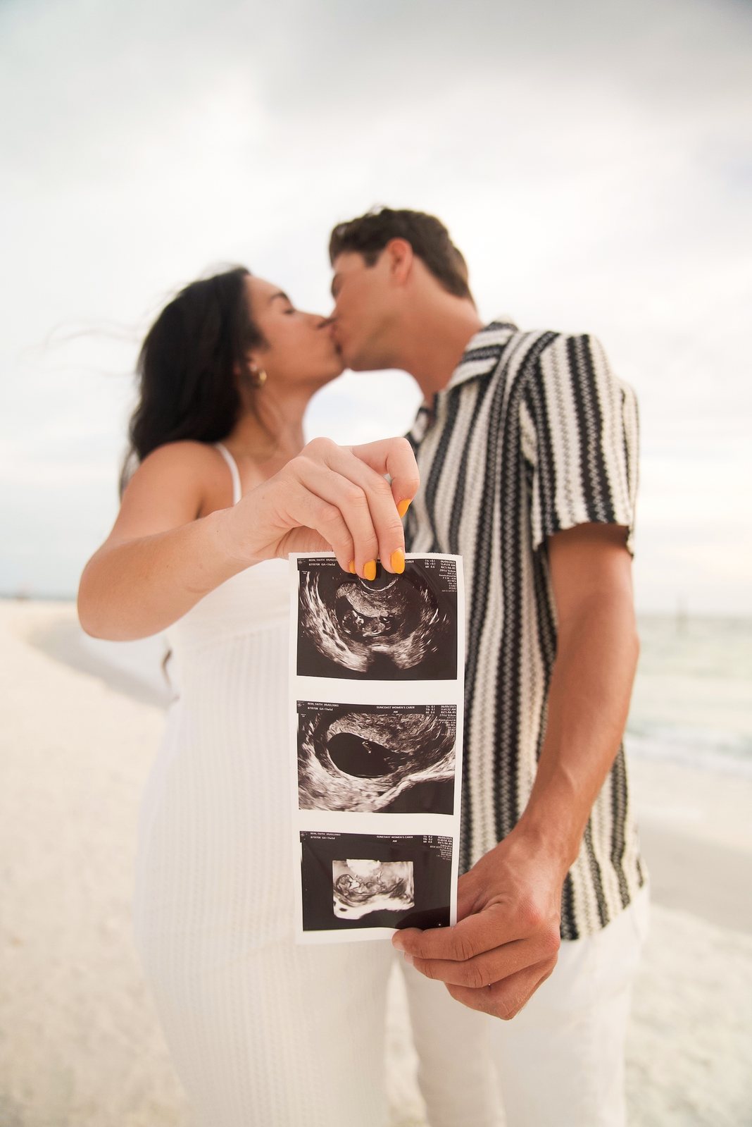 Couple kissing on the beach while holding up their baby announcement ultrasound