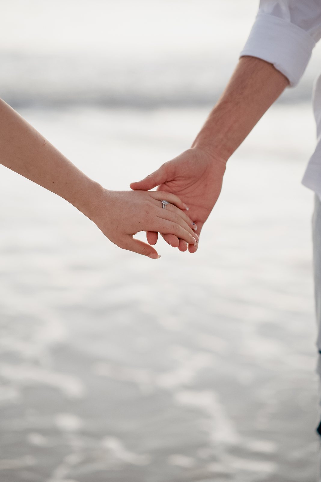 Couple holding hands on the beach, engagement ring in focus