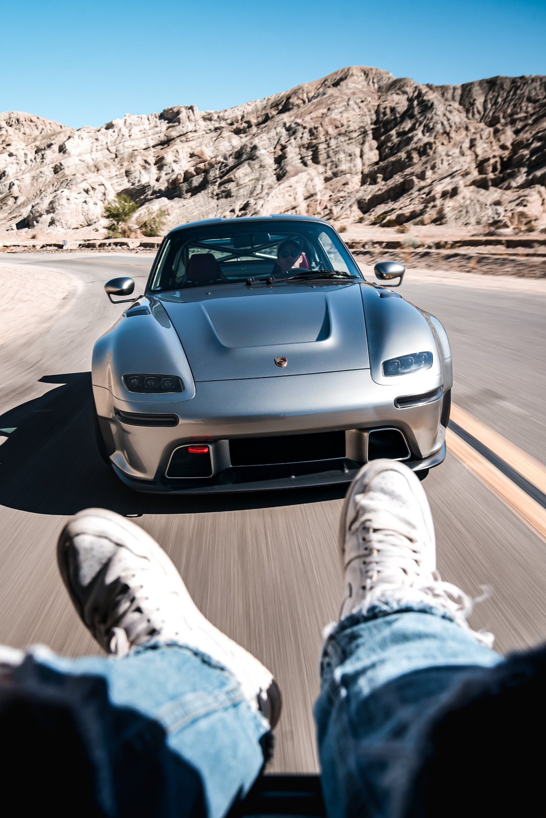 Silver restomod Porsche chasing on a desert canyon road, feet out the window