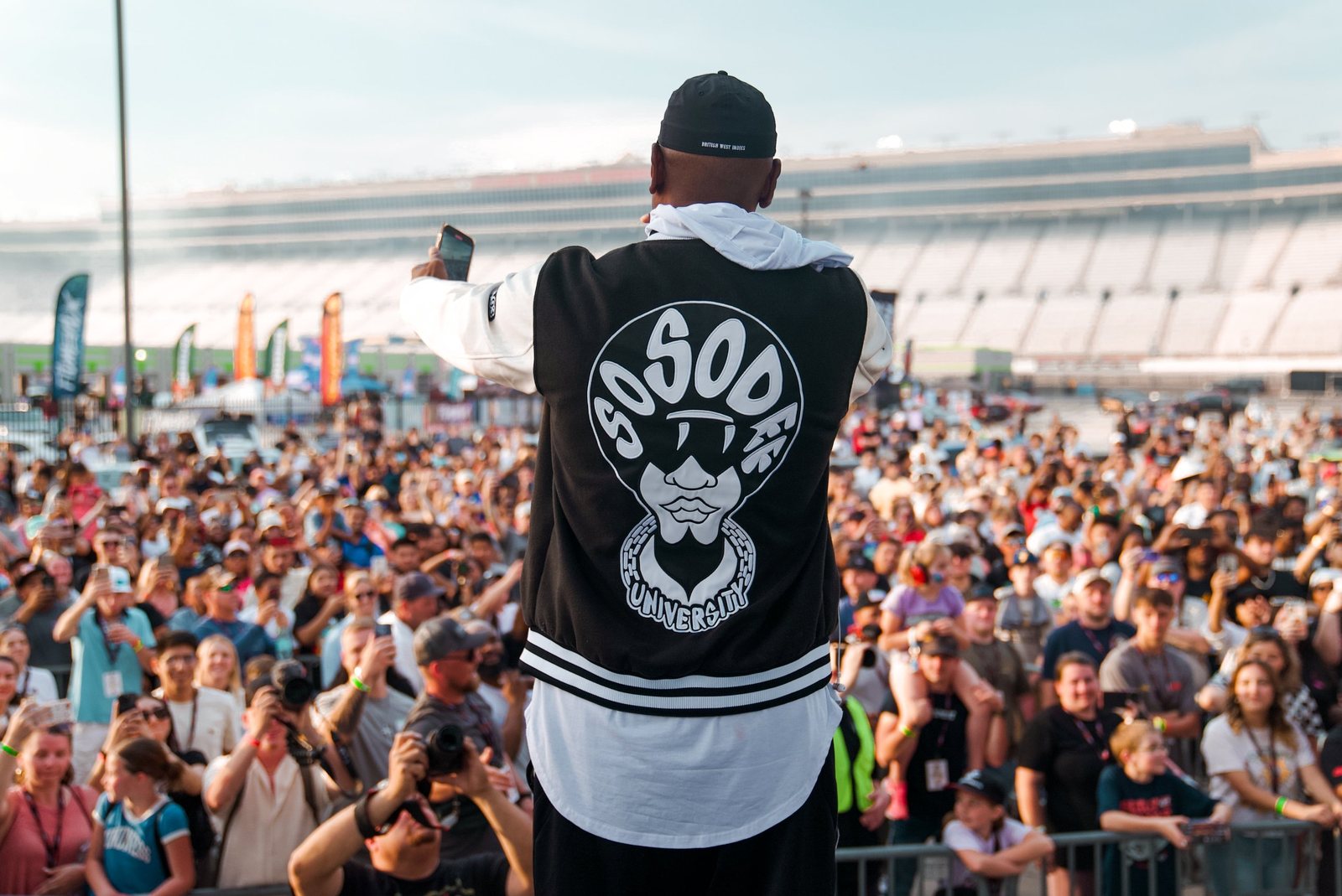 Performer in a So So Def varsity jacket filming the crowd from the stage at the speedway